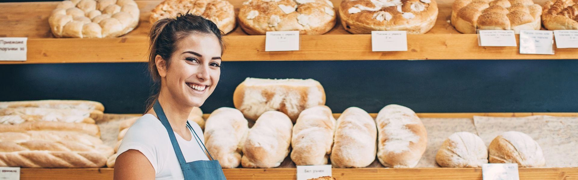 Mulher empreendedora sorrindo dentro de sua padaria e com uma vitrine de pães atrás dela.