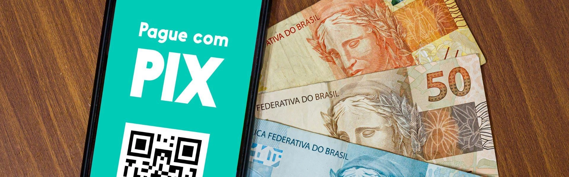 Na imagem temo um celular com "Pague com PIX" escrito na tela e ao lado dele, notas de reais.