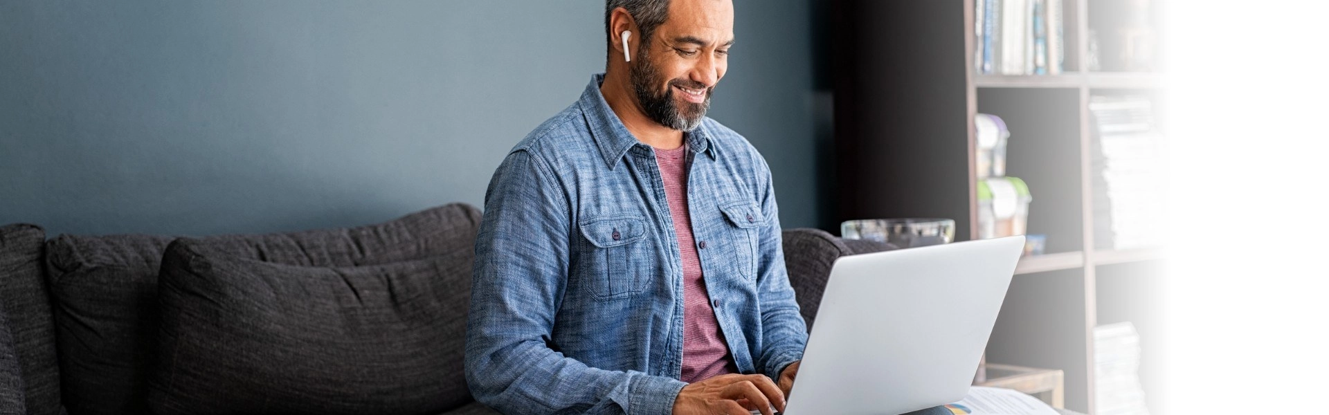 Homem sentado em um sofá, usando fones de ouvido e utilizando laptop enquanto sorri.