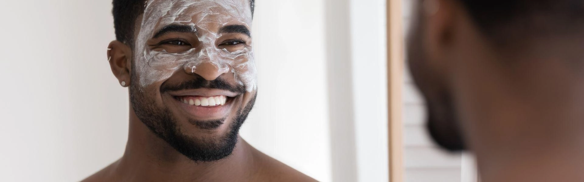 Homem negro sorrindo ao se olhar no espelho usando cosméticos masculinos.