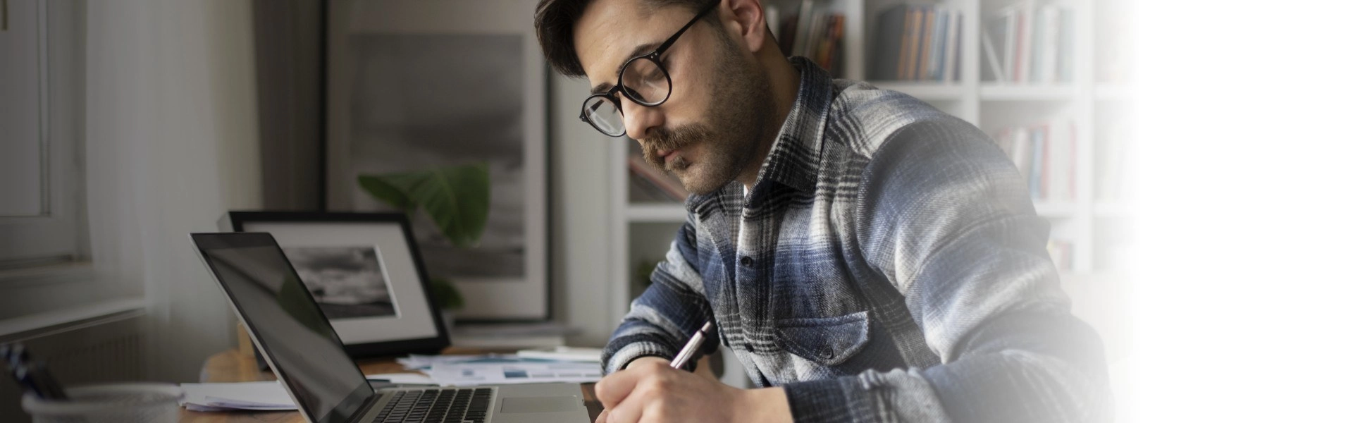 Homem branco de barba escreve com caneta ao lado de notebook em seu escritório