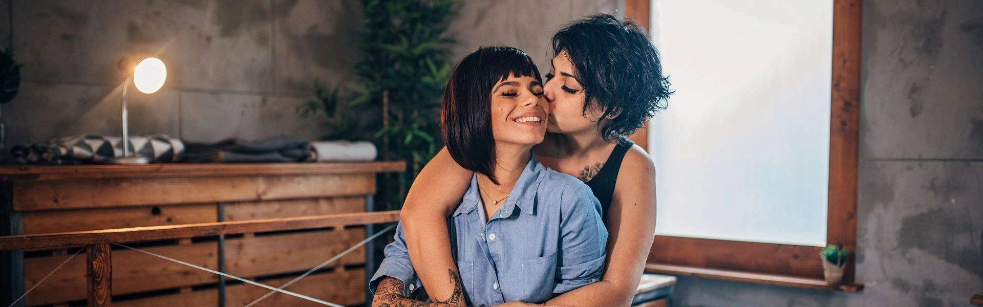 Casal de duas mulheres se abraçando em cima da cama, em um quarto.