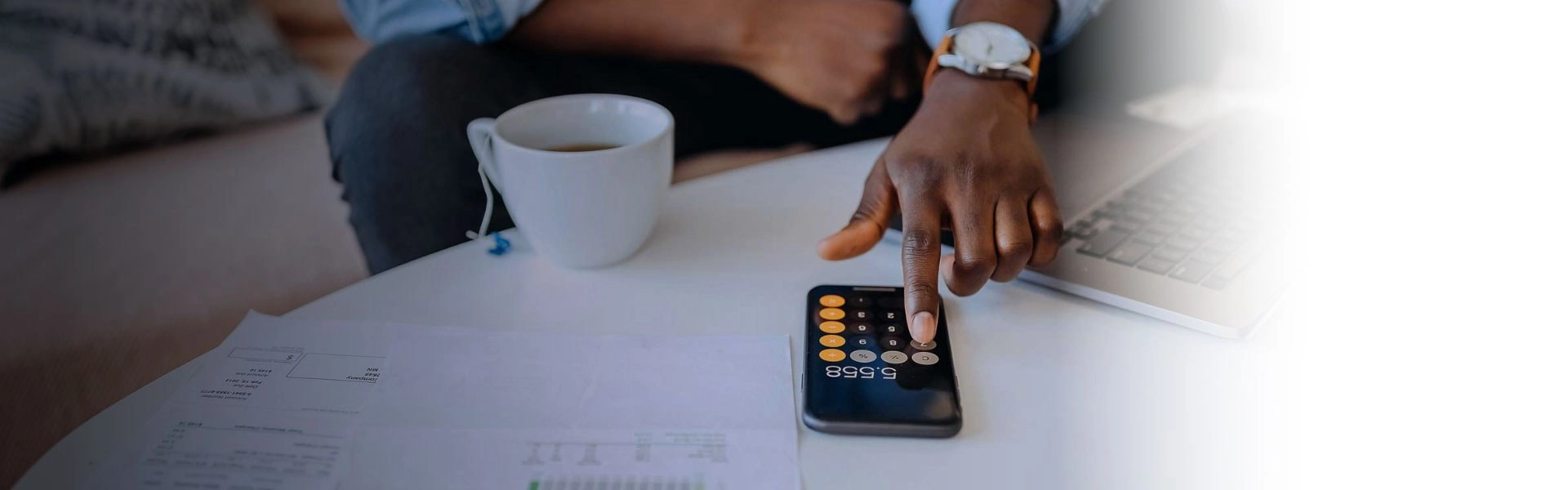 Mão de homem negro usando a calculadora no celular que está em cima de uma mesa branca com uma xícara de chá do lado.