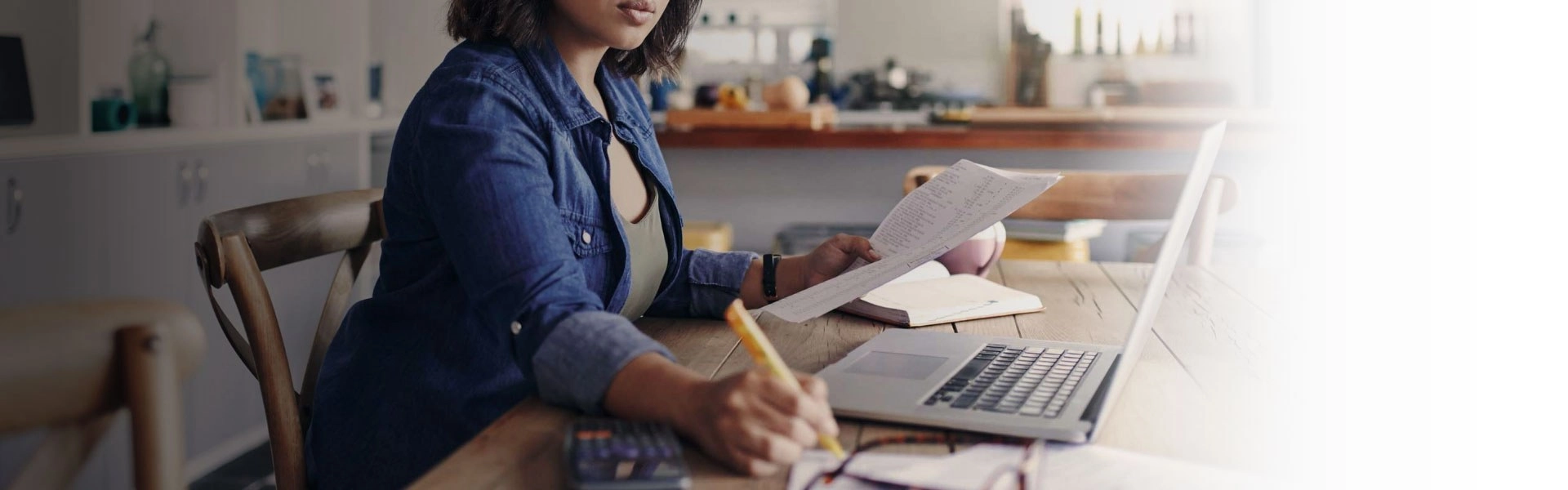 Mulher branca sentada com um notebook e uma calculadora em cima de uma mesa enquanto faz anotações com uma mão e segura uma folha na outra mão.