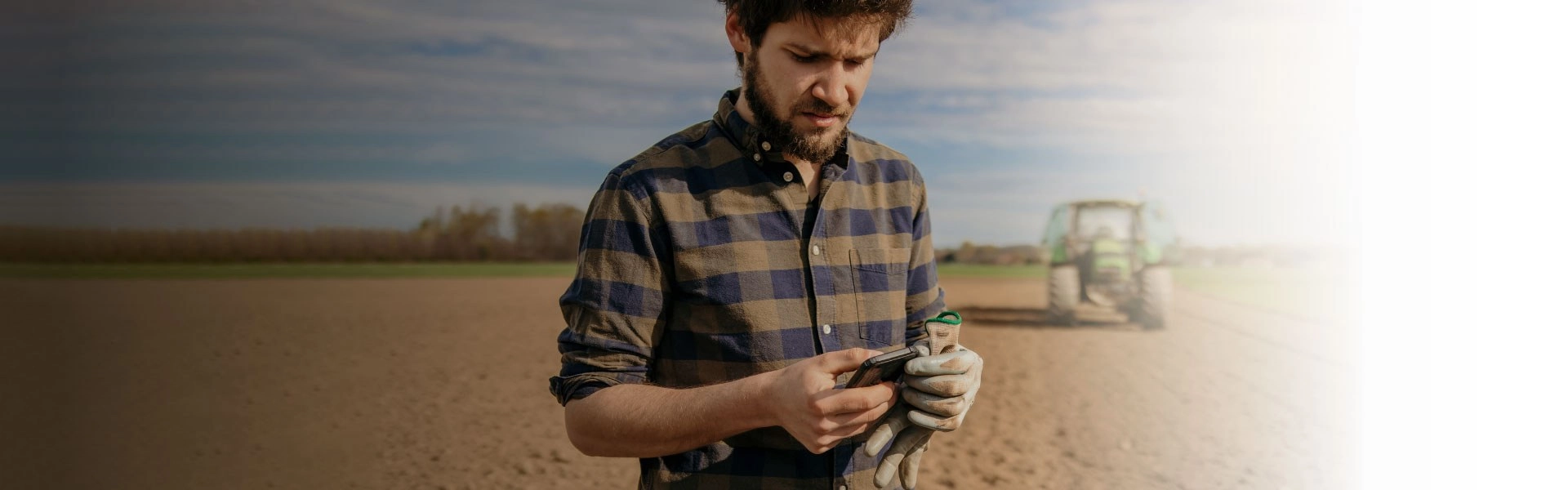 Homem branco no meio de um terreno de plantação mexendo no celular.