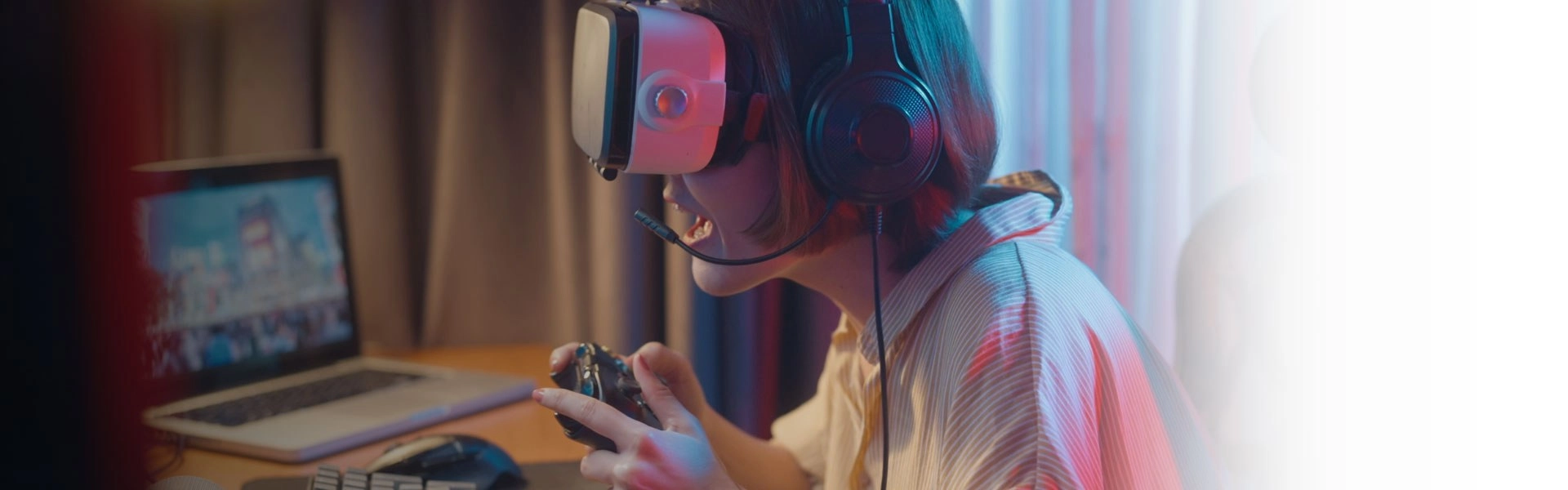Mulher branca usando óculos de realidade virtual e fones de ouvido segurando um controle de vídeo game em frente ao computador.