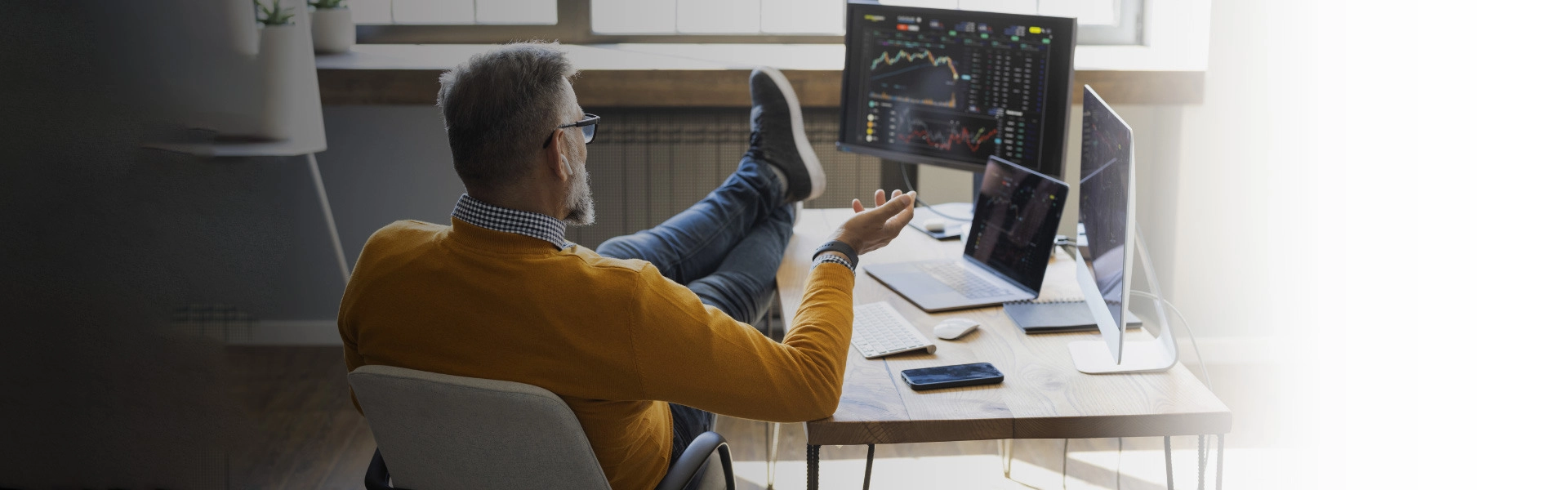 Homem branco sentado em uma cadeira com os pés apoiados em uma mesa com um notebook e dois monitores em cima.