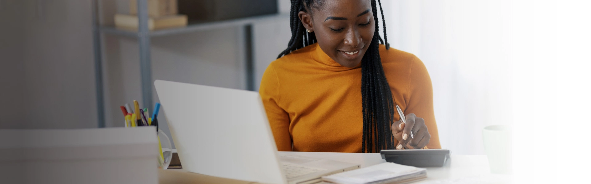 Mulher negra sorrindo sentada em frente a um notebook usando uma calculadora.