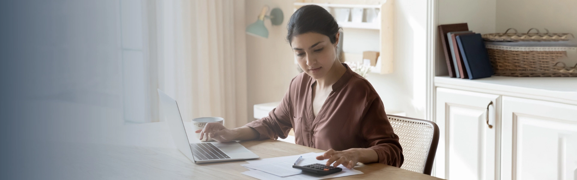 Mulher branca em uma mesa utilizando calculadora enquanto faz anotações em um laptop.