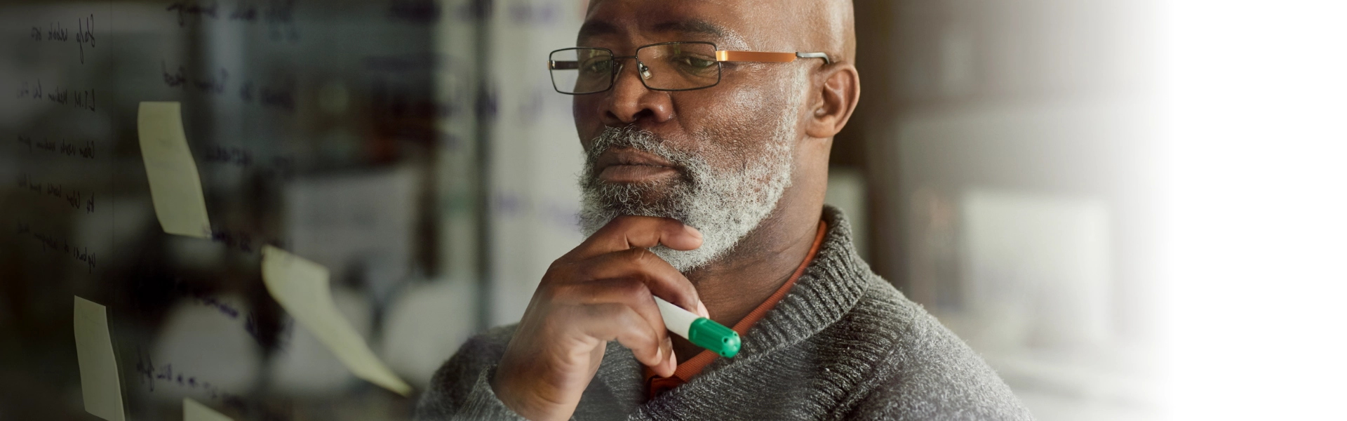 Homem negro com barba grisalha pensa sobre correr riscos segurando a caneta na mão direita. "