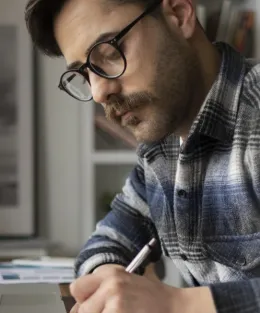 Homem branco de barba escreve com caneta ao lado de notebook em seu escritório