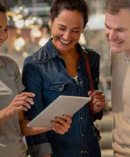 Uma vendedora branca mostra sorrindo um tablet para casal composto de homem e mulher brancos que sorriem também