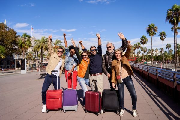 Grupo de amigos felizes se preparando para viajar, com malas ao fundo e palmeiras sob um céu azul claro, ideal para promover turismo e viagens em grupo.