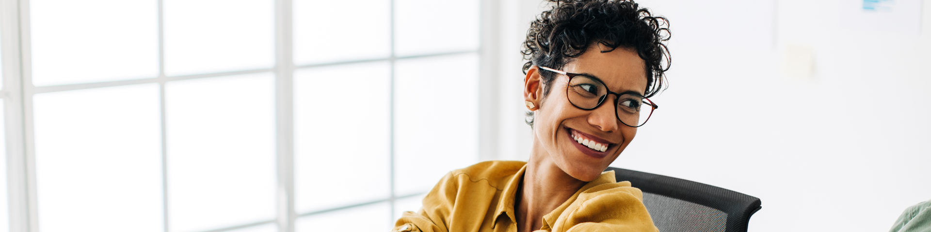 Mulher sorrindo, usando óculos e blazer amarelo, em ambiente de escritório com janelas grandes ao fundo.