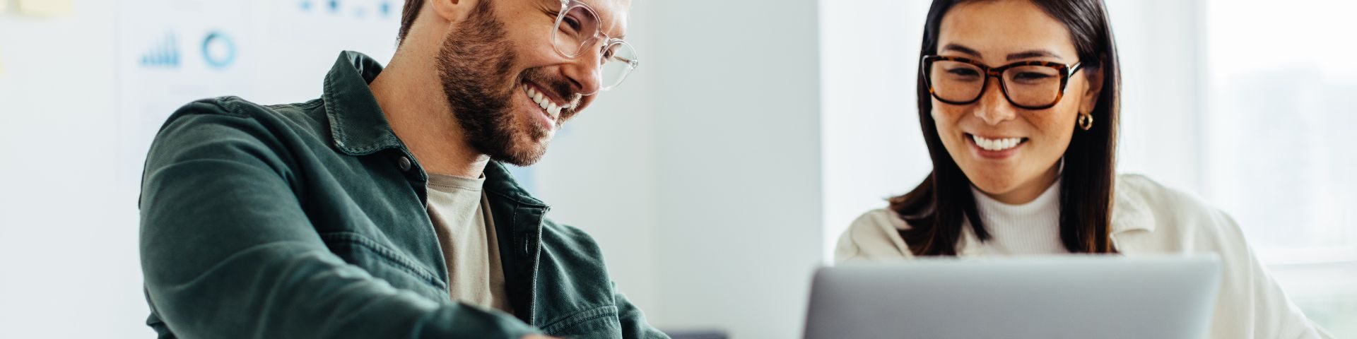 Homem e mulher sorrindo enquanto olham para um laptop, colaborando em um ambiente de escritório moderno, promovendo trabalho em equipe e inovação.