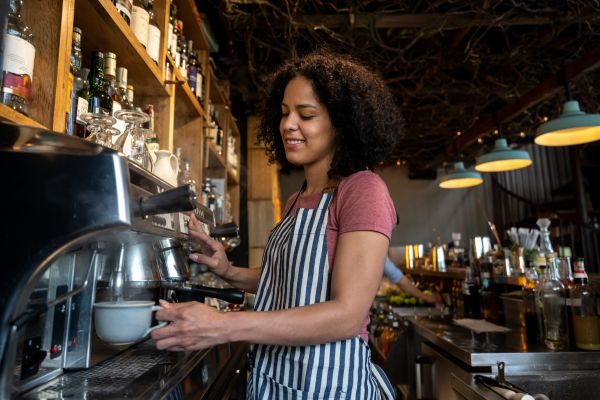 Mulher com avental, atrás do balcão de uma cafeteria, preparando café nas máquinas com expressão tranquila e feliz