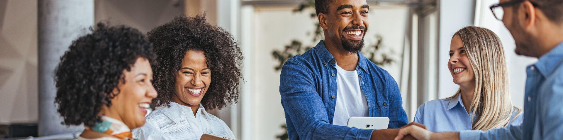 Grupo de profissionais diversos em um ambiente moderno de trabalho, sorrindo e cumprimentando-se de forma descontraída, transmitindo colaboração e networking.