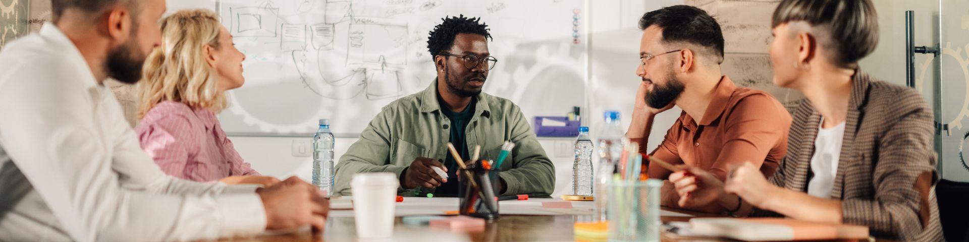 Equipe multicultural de profissionais reunida em torno de uma mesa, com materiais de apoio e quadro branco ao fundo, discutindo ideias para inovação e projetos.