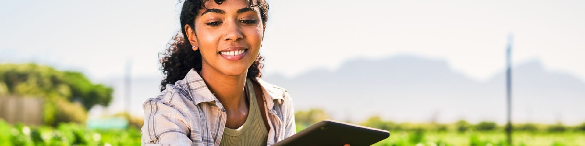 Mulher sorridente usando um tablet ao ar livre em ambiente rural, com vegetação e céu ao fundo, transmitindo alegria e conexão com a natureza.