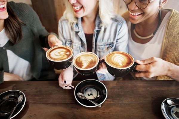 Três mulheres rindo e segurando xícaras de café, dando risada e compartilhando um momento de diversão juntas.