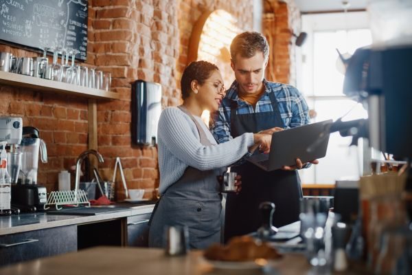 Homem e mulher atrás do balcão de uma cafeteria, mexendo em um computador enquanto conversam.