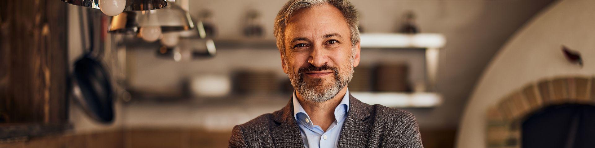 Homem de cabelo grisalho e barba bem cuidada sorrindo com ambiente acolhedor ao fundo, luz quente destaca sua expressão amigável.