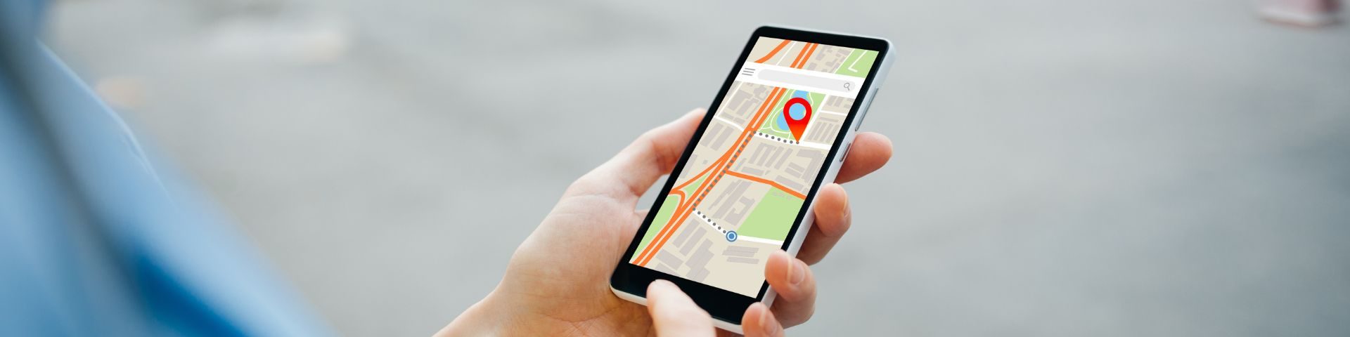 Pessoa usando um smartphone com um aplicativo de mapa na tela, segurando o dispositivo na mão, com fundo desfocado de rua ou calçada.