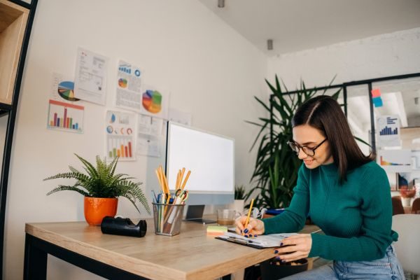 Mulher concentrada escrevendo em um caderno em um ambiente de trabalho moderno, cercada por plantas e materiais de escritório.