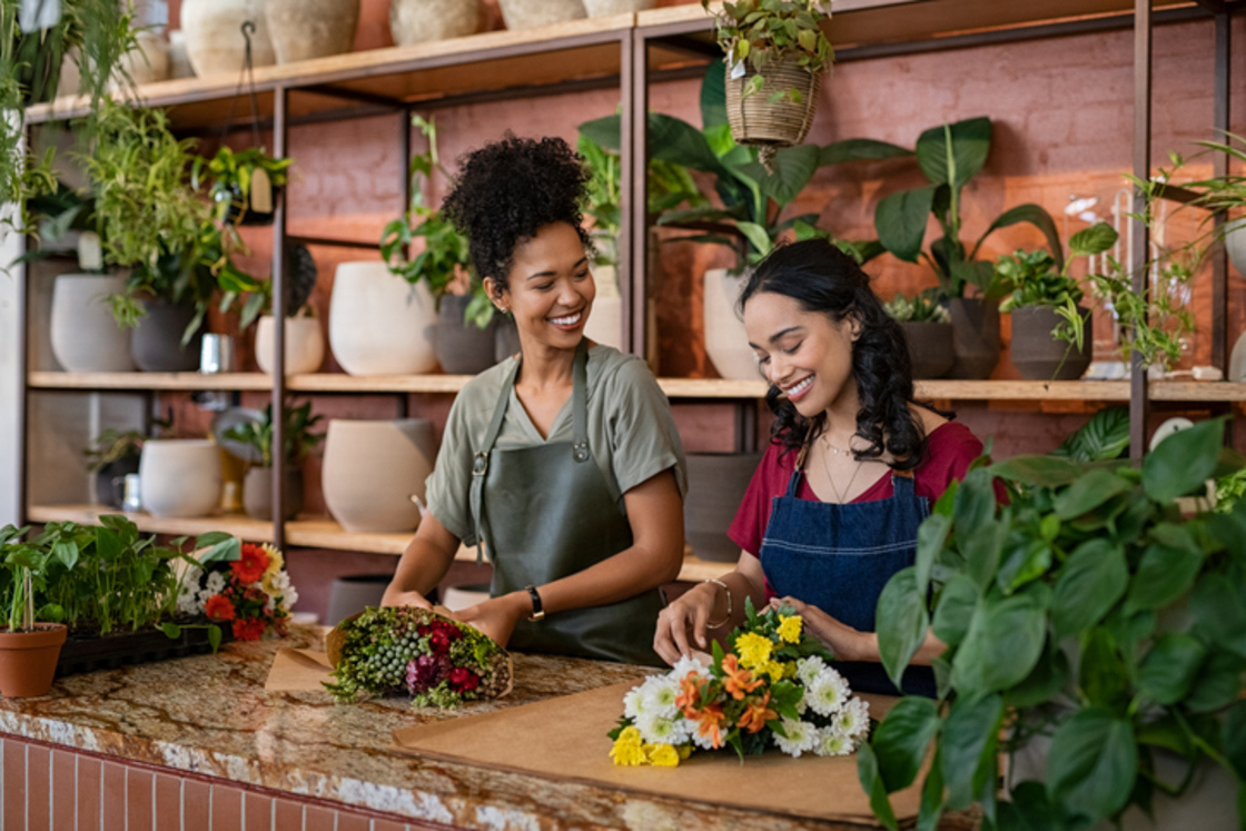 Duas mulheres sorrindo enquanto trabalham em uma floricultura, montando arranjos florais rodeadas por plantas em um ambiente acolhedor.