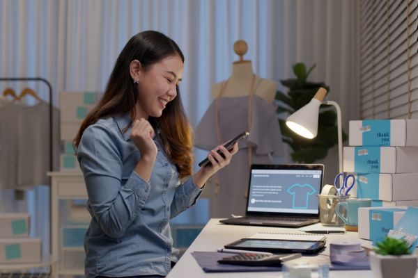 Mulher sorrindo enquanto utiliza um celular em um ambiente de escritório, cercada por materiais de trabalho e uma tela de computador com gráficos.