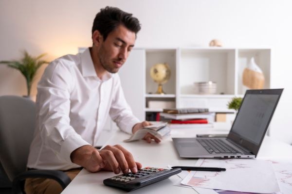 Homem trabalhando em um escritório moderno, usando uma calculadora e analisando documentos. A mesa possui um laptop e materiais de escritório.