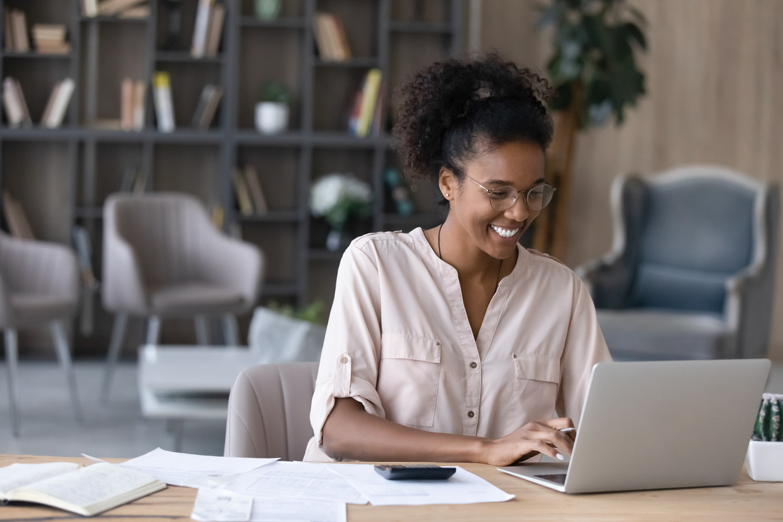 Mulher sorridente trabalhando em um laptop em um ambiente moderno e aconchegante, representando um estilo de vida produtivo e equilibrado.