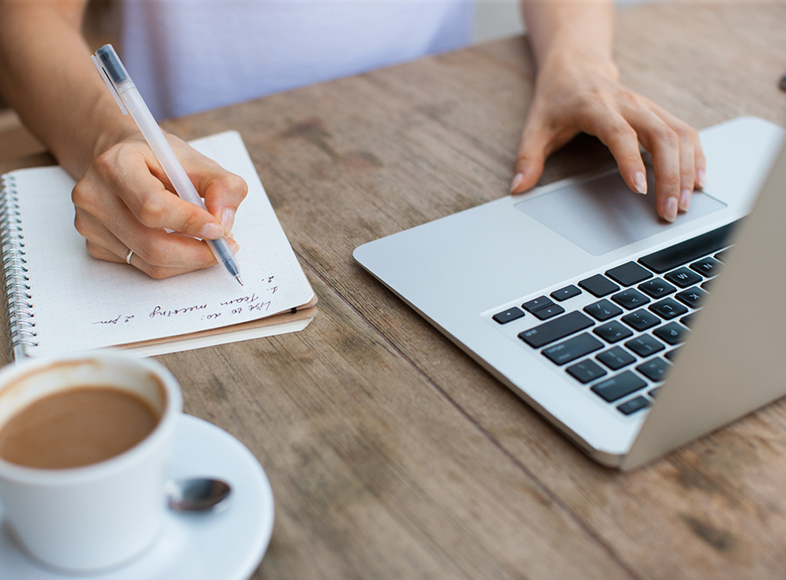 Pessoa escrevendo em um caderno enquanto utiliza um laptop em uma mesa de madeira com uma xícara de café ao lado, ideal para inspirar produtividade e foco.