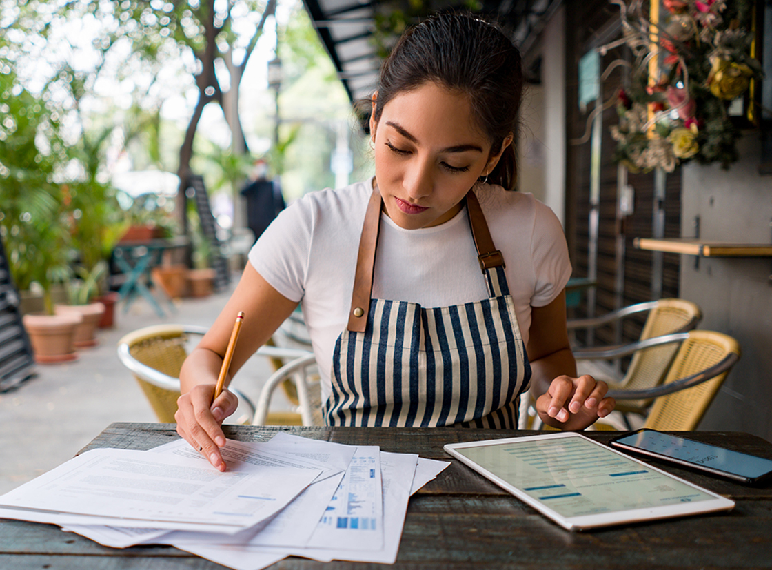 Mulher trabalhando em um café, revisando documentos e usando tablet em ambiente agradável e tranquilo.