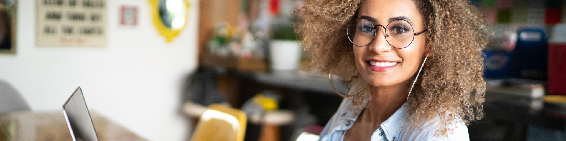 Jovem mulher de cabelo cacheado, usando óculos e fone de ouvido, trabalhando e sorrindo ao lado de um laptop em um espaço de coworking moderno.