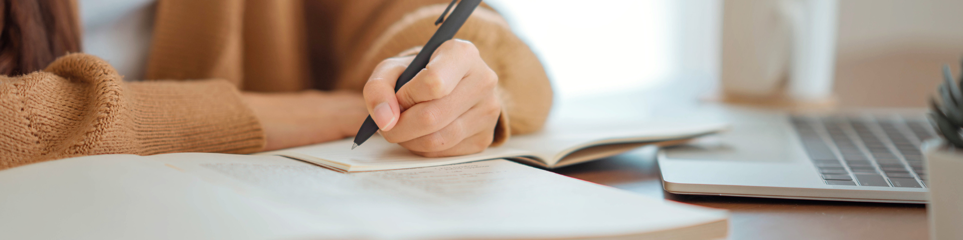 Jovem estudando e fazendo anotações em um caderno, com laptop e caneta, em um ambiente bem iluminado, ideal para estudos e produtividade.