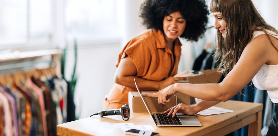 Duas mulheres interagindo em um ambiente de trabalho, uma usando um laptop enquanto a outra observa, simbolizando colaboração e tecnologia no comércio.