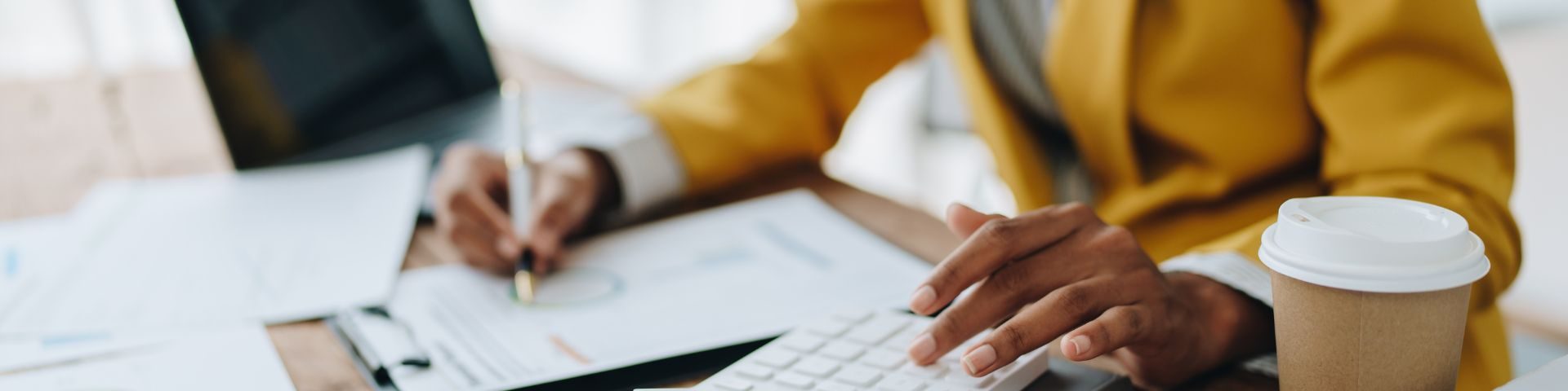 Pessoa trabalhando em um laptop com documentos e uma xícara de café em um ambiente de escritório.