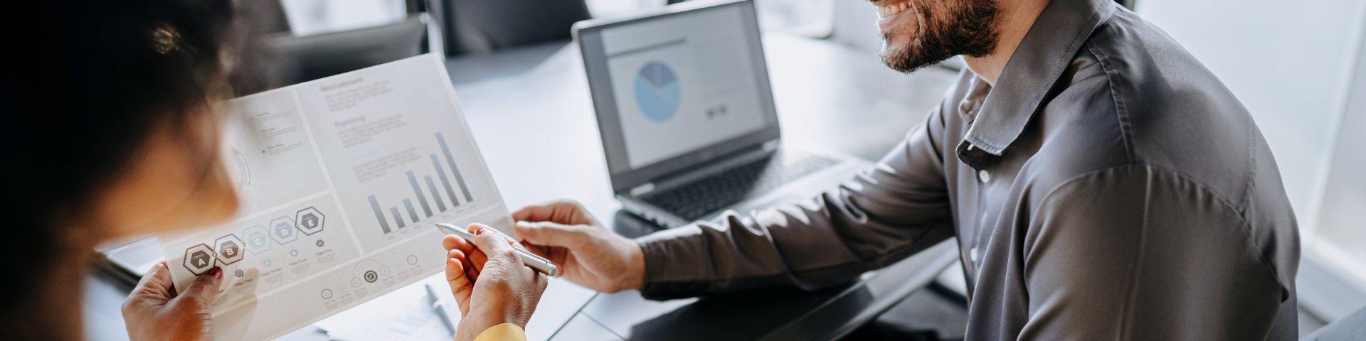 Reunião de negócios com duas pessoas discutindo gráficos e dados em escritório moderno, representando estratégia empresarial e análise de resultados.