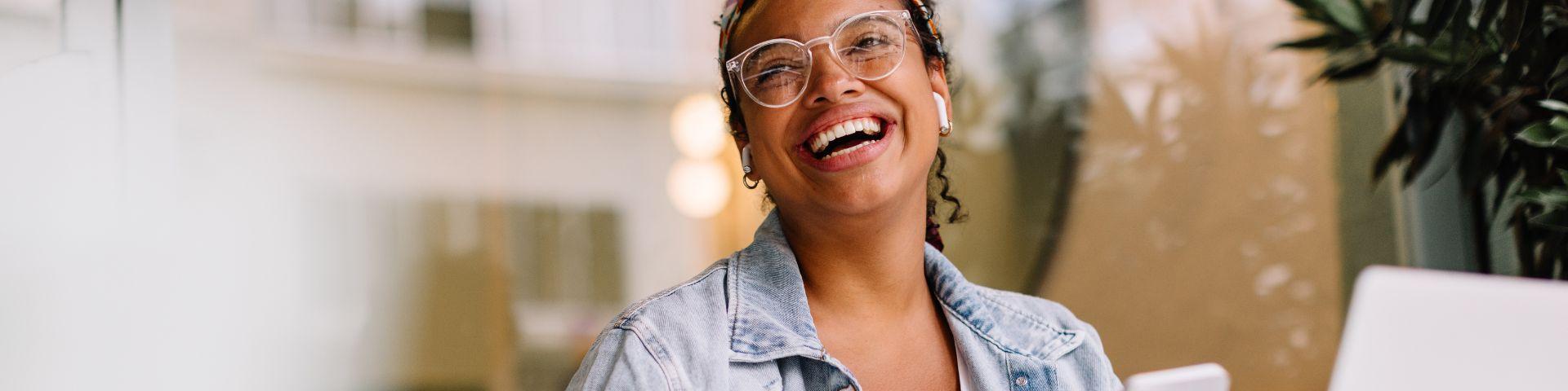 Mulher sorridente utiliza notebook e celular em uma cafeteria, segurando cartão de crédito, representando o acesso facilitado a soluções financeiras digitais.