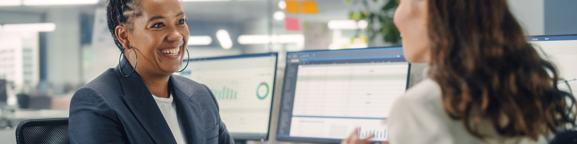 Mulher empreendedora em reunião corporativa, sorridente, discute indicadores financeiros exibidos em telas de computador, ilustrando planejamento e gestão de crédito para negócios.