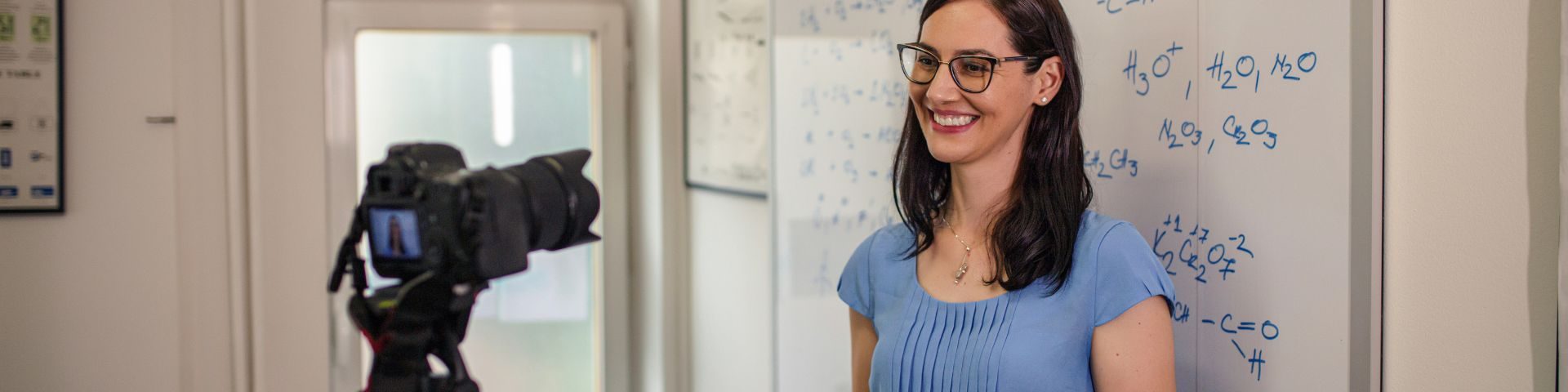 Mulher com óculos e blusa azul sorri enquanto é filmada por uma câmera profissional diante de um quadro branco com fórmulas químicas escritas em azul, durante a gravação de uma aula de curso livre.