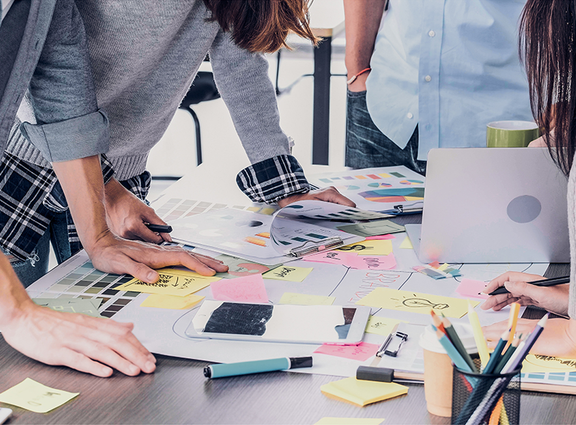 Grupo de pessoas colaborando em um ambiente de trabalho, analisando esboços e anotações coloridas sobre uma mesa.