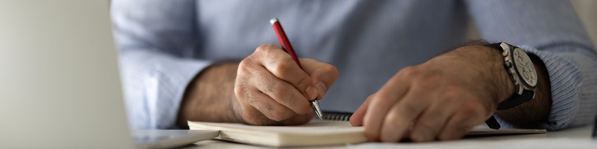 Pessoa escrevendo em um caderno com uma caneta vermelha, usando um relógio de pulso e vestindo uma camisa cinza, demonstrando foco e produtividade.