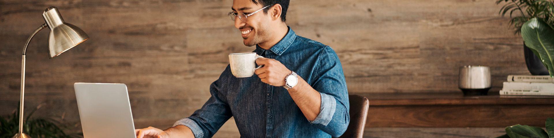 Homem sorridente tomando café enquanto trabalha em um computador portátil em um ambiente aconchegante de escritório com decoração moderna e plantas ao fundo.