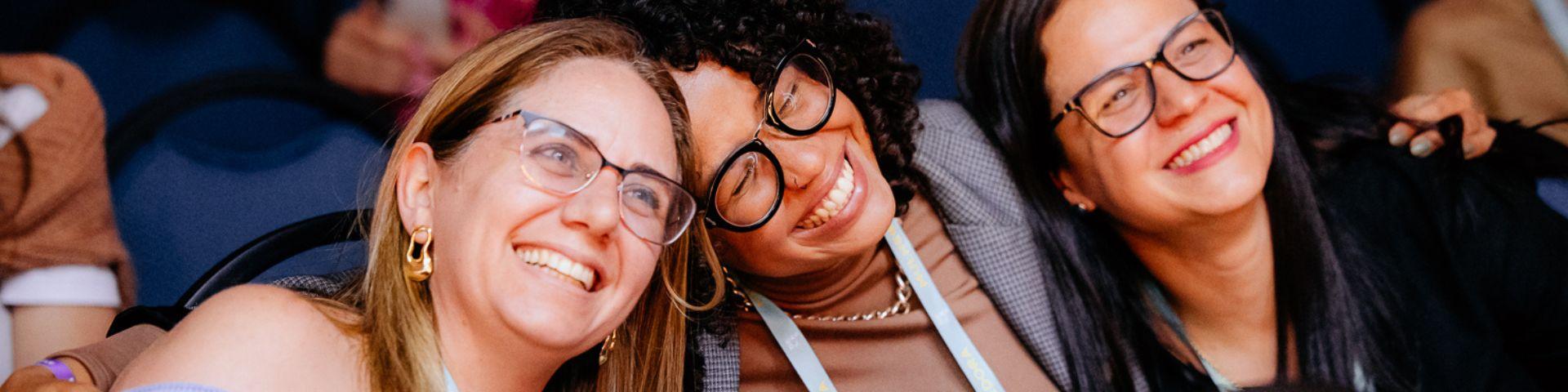 Três mulheres sorridentes usando óculos e apresentando diversidade de etnias, participando do Delas Summit, em um ambiente com cadeiras azuis.