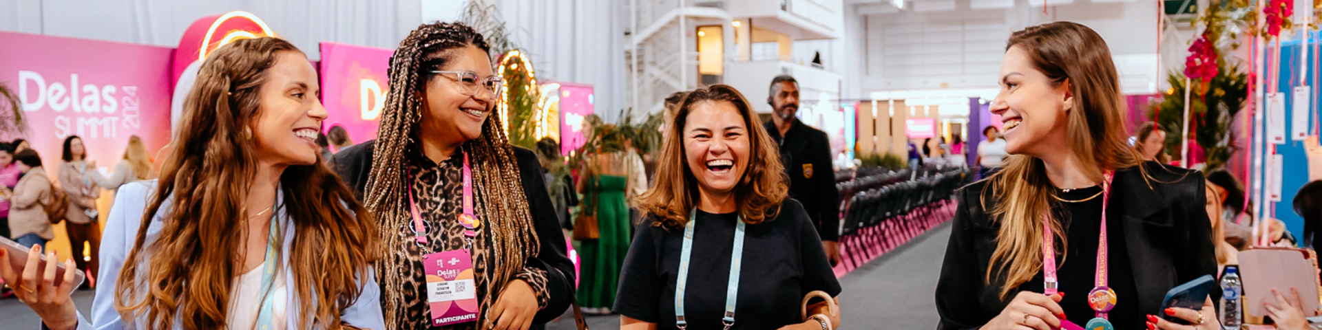 Quatro mulheres sorrindo e conversando enquanto caminham no Delas Summit, usando crachás coloridos e rodeadas por estandes decorados em tons de rosa.