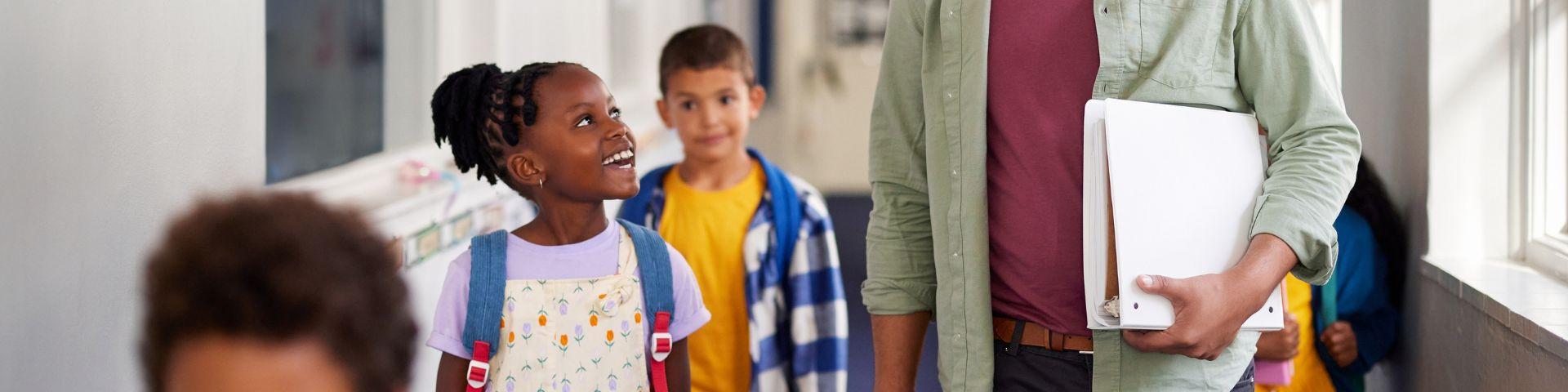 Crianças e professor conversando em corredor escolar, com foco na interação alegre entre educador e estudantes em ambiente de escola.