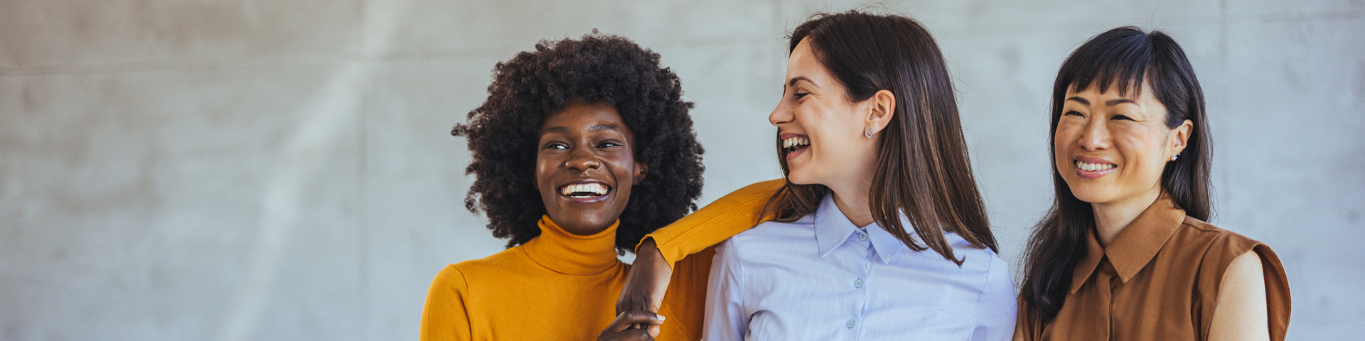 Três mulheres sorrindo lado a lado; uma veste blusa amarela, outra camisa azul-clara e a terceira blusa marrom, em um ambiente interno iluminado.