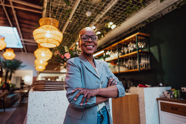 Mulher de sorriso acolhedor em um ambiente de restaurante moderno, vestindo um blazer claro. O local tem iluminação suave e decoração elegante, ideal para confraternizações.